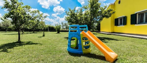 Un parque infantil con tobogán y estructuras para escalar en un área verde con árboles y un edificio amarillo al fondo.