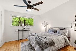 A bedroom with a bed, a ceiling fan, a window with a view of trees, a bedside table, and a wall-mounted lamp. - World Cup Villa · 7-Min Walk to AT&T Stadium (Arlington)