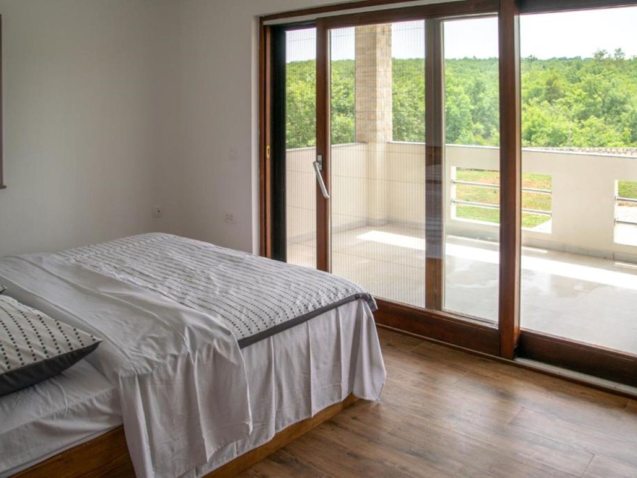 A bedroom with a bed, a sliding glass door leading to a balcony, and a view of greenery outside.