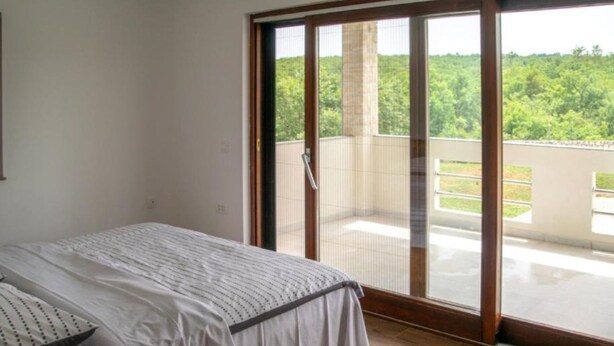 Quarto com cama, porta de correr de vidro que dá para uma varanda e vista para o verde lá fora.