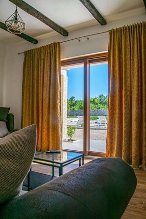 Ein Zimmer mit einer Couch, einem Glastisch und Vorhängen. Blick auf einen Außenbereich mit einer Steinmauer und viel Grün.