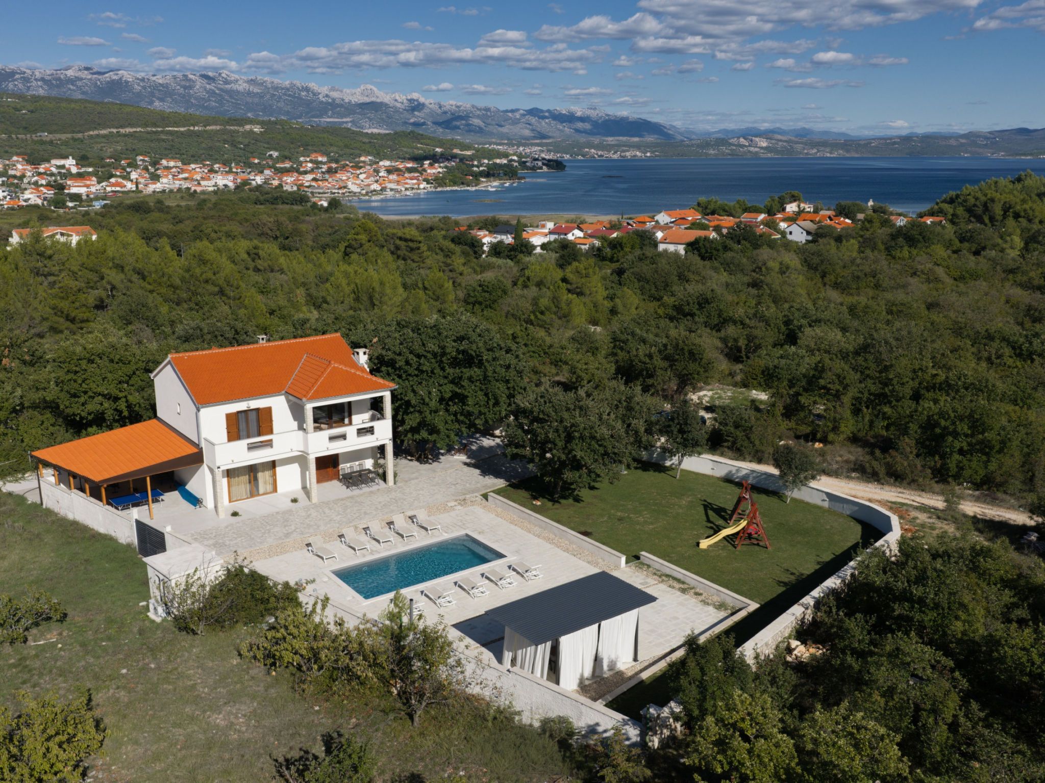 A modern two-story house with a swimming pool, surrounded by lush greenery and a view of a coastal town.