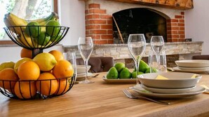 Uma mesa de jantar com frutueiras, copos de vinho e pratos, uma lareira de tijolos e vista para árvores lá fora.