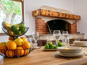 Ein Esstisch mit Obstschalen, Weingläsern und Tellern, ein Kamin aus Ziegelsteinen und ein Blick auf Bäume draußen.