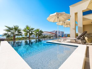 A swimming pool with lounge chairs and umbrellas, surrounded by palm trees and buildings. - Villa Novue with pool by Interhome (Rovanjska)