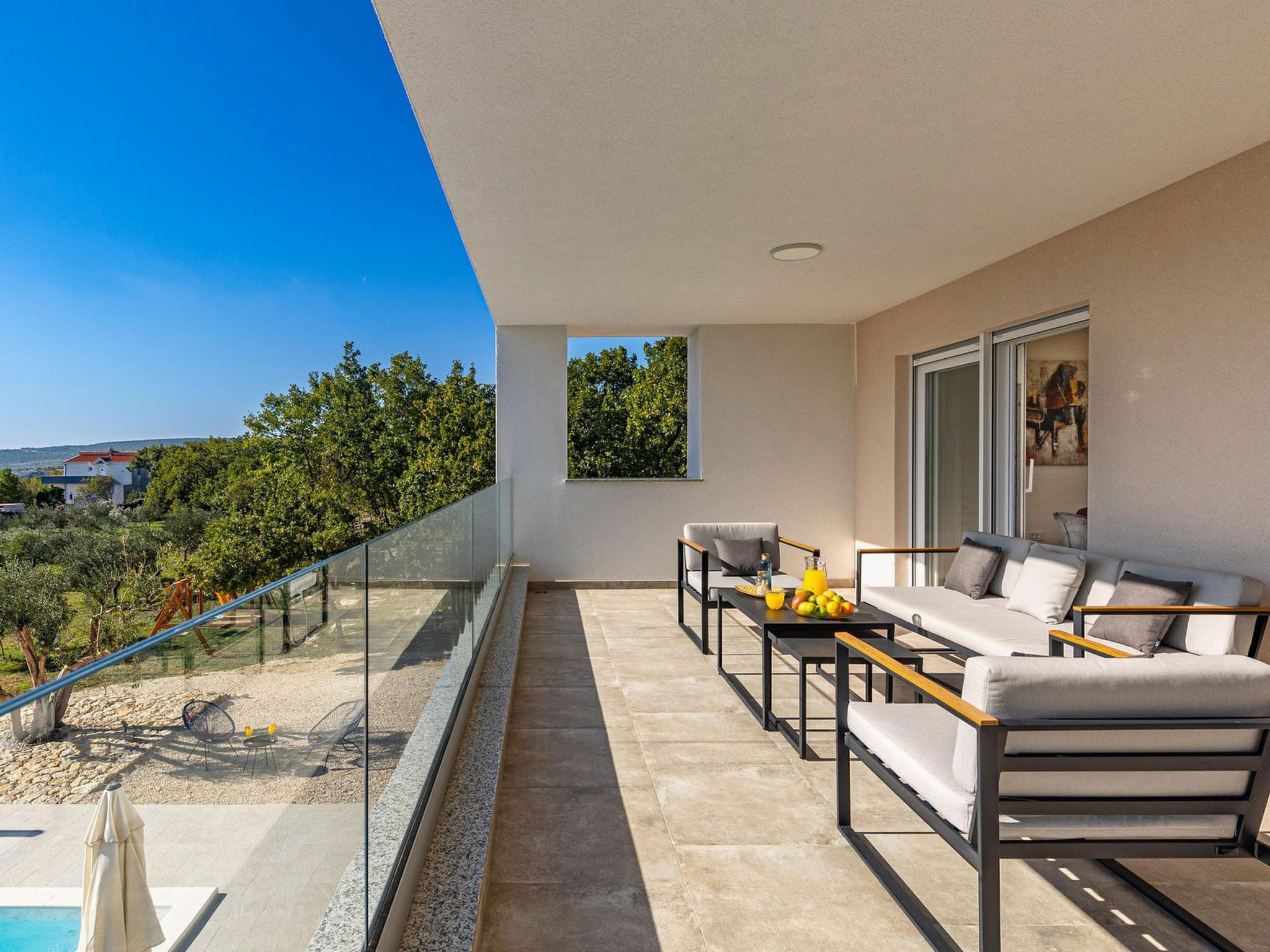 A balcony with a glass railing, a sofa, a table with fruit, and a view of the landscape.