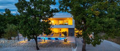 A modern two-story building with a swimming pool, outdoor seating, and trees.
