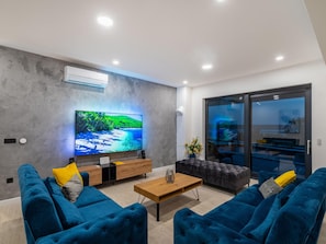 Uma sala de estar moderna com uma televisão de tela grande, uma mesa de centro de madeira e sofás azuis.