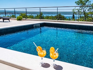 Uma piscina com água azul cristalina, duas bebidas com fatias de laranja, e vista para o mar e colinas ao fundo.