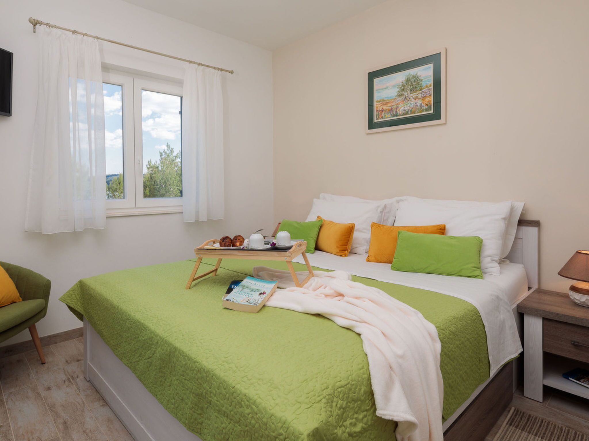 Una cama bien hecha con una manta verde, almohadas en tonos verde y naranja, una mesita de noche con una lámpara y una ventana con cortinas blancas.