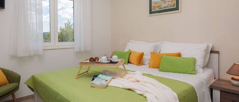 A neatly made bed with a green blanket, pillows in green and orange, a bedside table with a lamp, and a window with white curtains.
