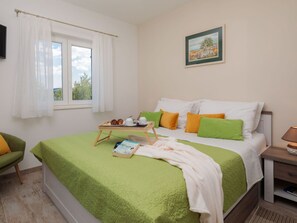Ein ordentlich bezogenes Bett mit einer grünen Decke, Kissen in Grün und Orange, ein Nachttisch mit einer Lampe und ein Fenster mit weißen Vorhängen.