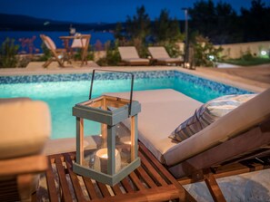 A poolside area with lounge chairs, a wooden table with a lantern, and a view of the pool. - Vacation home  by Interhome (Pridraga)