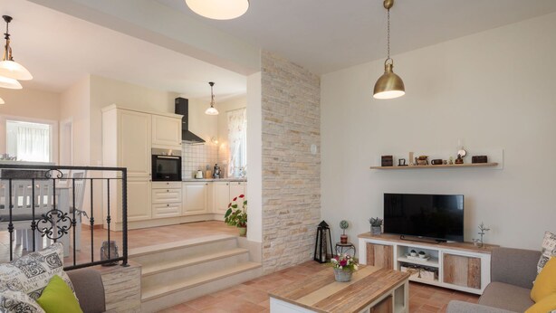 Un salon moderne avec un canapé, une table basse et un téléviseur. On aperçoit une cuisine à l’arrière-plan, avec des meubles blancs et un mur d’accent en pierre.