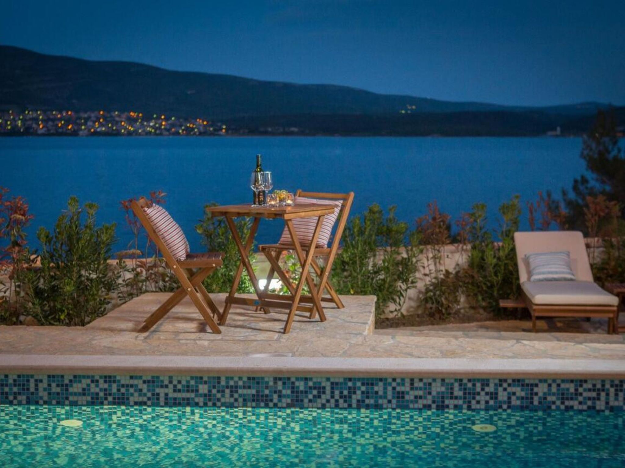 A poolside setting with a table and chairs, a lounge chair, and a view of a body of water with distant lights.
