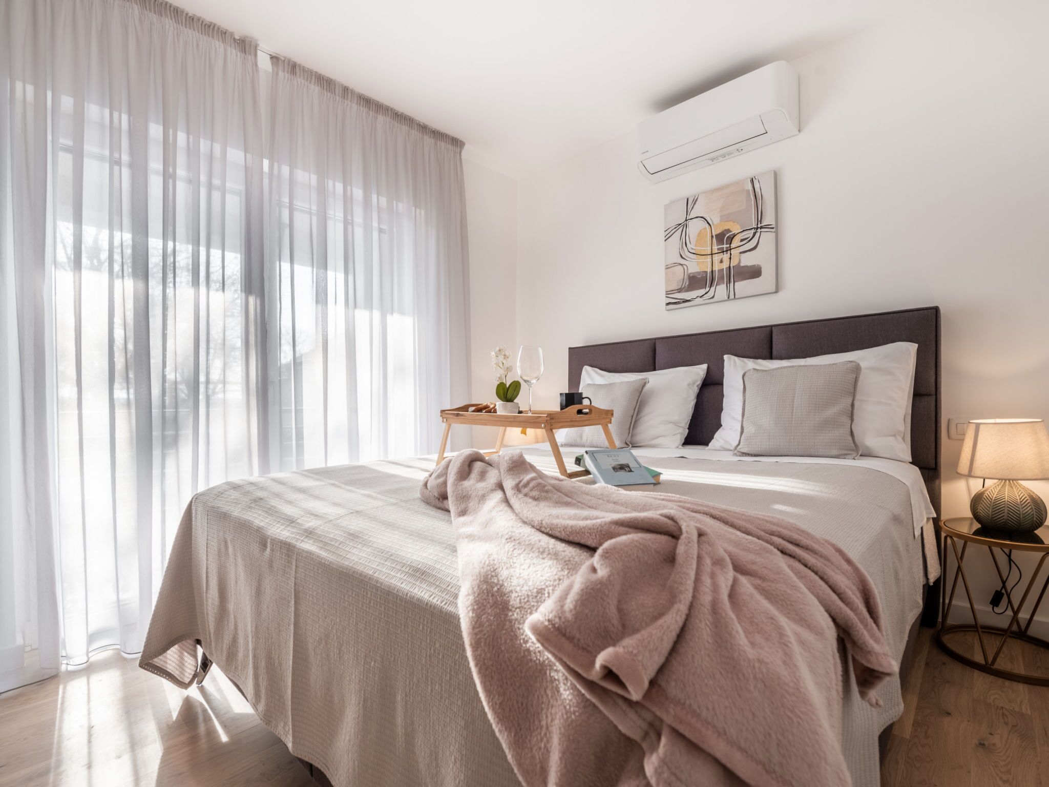 A modern bedroom with a large bed, a bedside table, a lamp, and a window with sheer curtains.