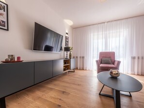 Ein modernes Wohnzimmer mit flachbildfernseher an der Wand, einem grauen Sofa, einem runden Couchtisch mit dekorativer Schale und einem großen Fenster mit transparenten Vorhängen.