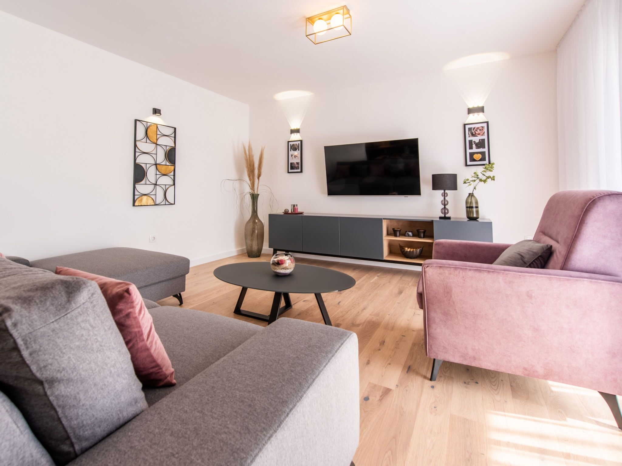 Una sala de estar moderna con un sofá gris, un sillón rosa, una mesa de centro redonda y un televisor de pantalla plana montado en la pared.