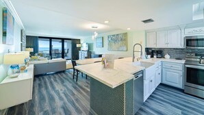 A modern kitchen with a central island, a dining area with a sofa, and a view of the ocean through large windows. - Beautifully Decorated 2BR 2BA BEACHFRONT Condo! (Miramar Beach)
