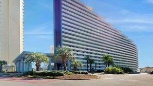 A modern hotel with a curved facade, a prominent vertical entrance, and a landscaped courtyard with palm trees. - Beautifully Decorated 2BR 2BA BEACHFRONT Condo! (Miramar Beach)
