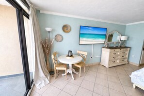 A room with a round table, two chairs, a dresser, a painting of a beach, and a window with curtains. - Oceanfront Studio | Pools + Sleeps 4 (Myrtle Beach)