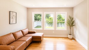 A room with a brown leather sofa, three windows, a potted plant, and a framed picture on the wall. - Welcome to Seven Oaks:Your Home Away From Home (City of Orange)