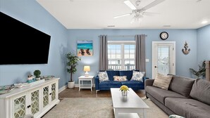 Ein Wohnzimmer mit blauem Sofa, weißem Couchtisch, grauem Sofa, Wandfernseher und Deckenventilator.