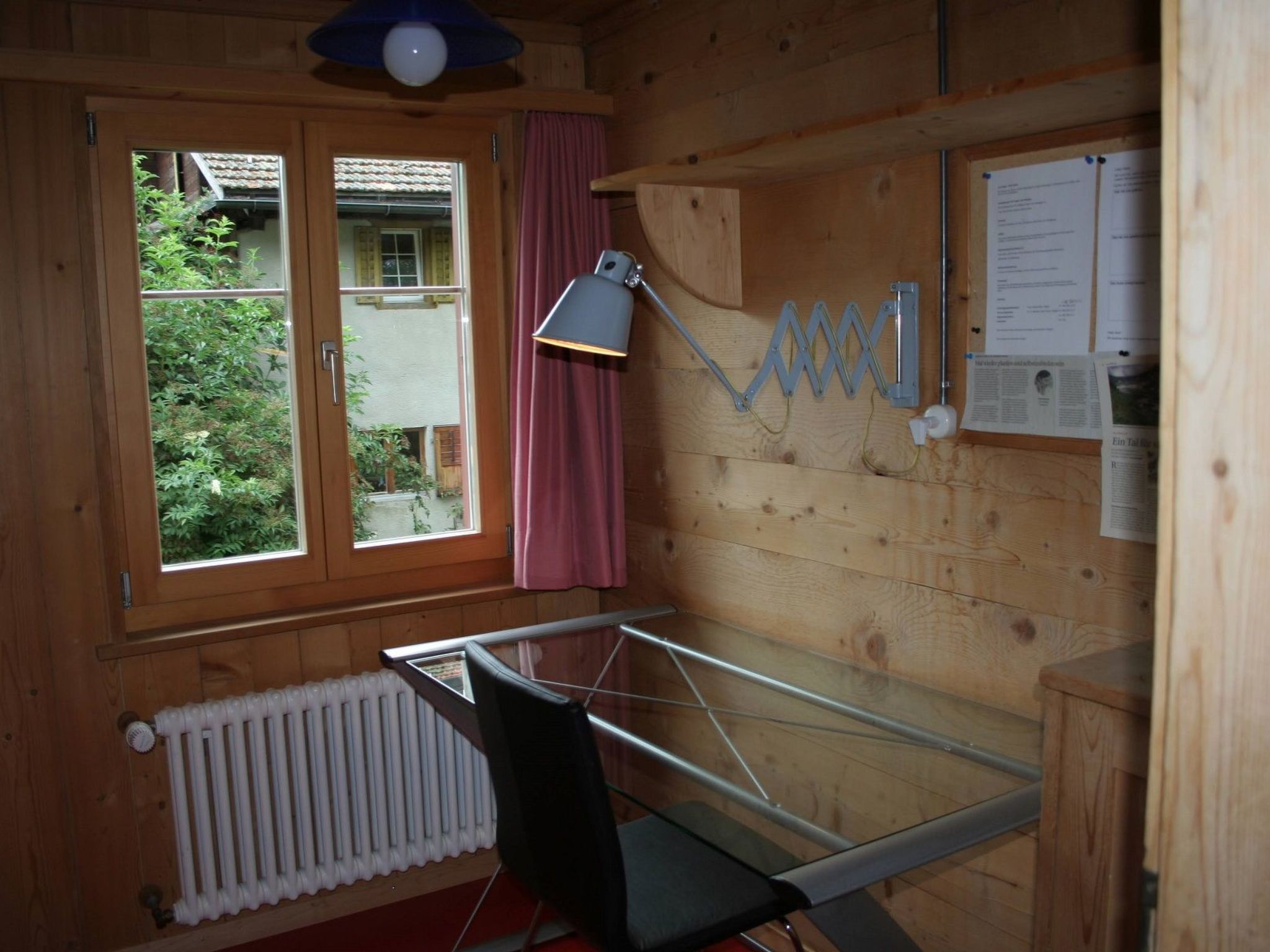 Ein Zimmer mit Holzvertäfelung, einem fenster mit Vorhängen, einem Heizkörper, einem Schreibtisch mit Stuhl, einer Schreibtischlampe und einem an der Wand befestigten Regal.