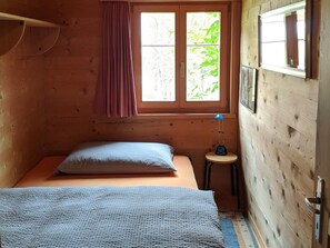 Una habitación pequeña de madera con una cama individual, una ventana con cortinas y un taburete.