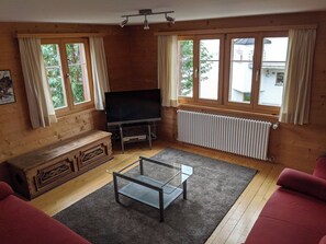 Ein Zimmer mit Holzvertäfelung, einem Fernseher auf einem Standfuß, einer Couch, einem Glastisch und Fenstern mit Vorhängen.