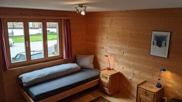 Ein schlichter Raum mit einem Einzelbett, einem Nachttisch, einem Schreibtisch und einem Fenster mit Vorhängen.