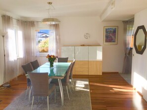 Ein Essbereich mit einem Glastisch, Stühlen, einer Blumenvase und Blick auf ein Haus durch das Fenster.