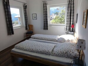 Quarto com duas camas, uma cadeira, uma mesa, um abajur e uma janela com cortinas.