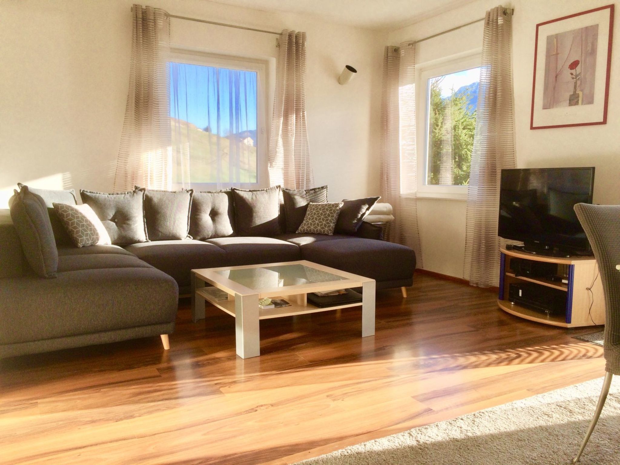 Una sala de estar con un sofá seccional, una mesa de centro, un televisor y un amplio ventanal con cortinas.