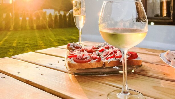 Une table en bois, sur laquelle se trouvent un verre de vin blanc et une assiette de nourriture, est placée à l’extérieur, avec une maison en arrière-plan.