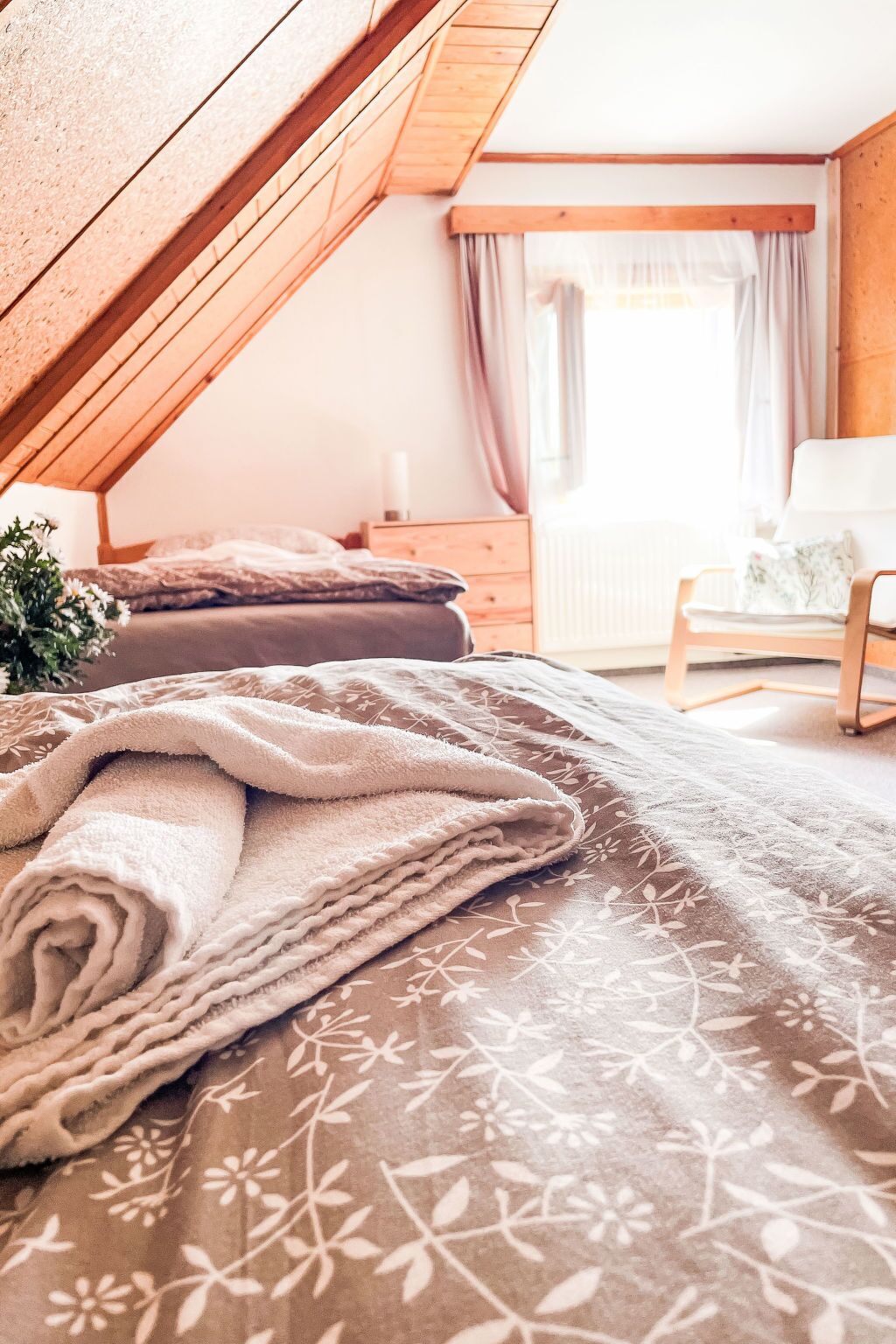 Une chambre avec un plafond en pente, un lit recouvert d’une couvre-lit à motifs floraux, une table de chevet, une chaise et une fenêtre avec des rideaux.