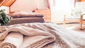 Une chambre avec un plafond en pente, un lit recouvert d’une couvre-lit à motifs floraux, une table de chevet, une chaise et une fenêtre avec des rideaux.