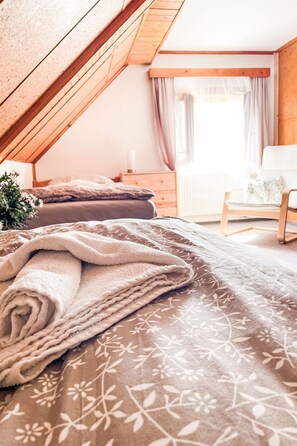 Ein Schlafzimmer mit geneigter Decke, einem Bett mit blumigem Überwurf, einem Nachttisch, einem Stuhl und einem Fenster mit Vorhängen.