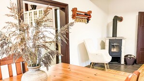 Une table à manger en bois, ornée d’un vase contenant des plantes séchées, une chaise blanche, un banc en bois et un poêle à bois.