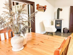 Ein Holztisch mit einer Vase gefüllt mit getrockneten Pflanzen, ein weißer Stuhl, eine Holzbank und ein Kaminofen.