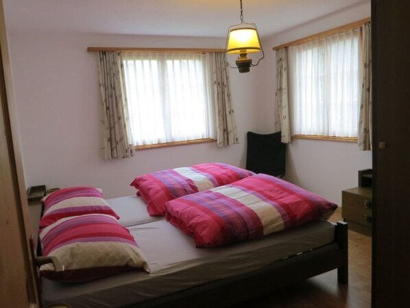 Un dormitorio con cama, dos almohadas, una mesita de noche, una lámpara y dos ventanas con cortinas.