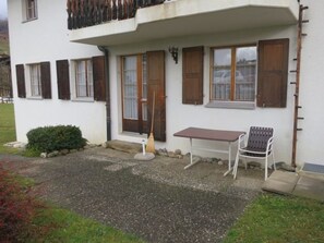 Un edificio de dos plantas con un camino de grava, un pequeño patio con una mesa y una silla, y una valla de madera.