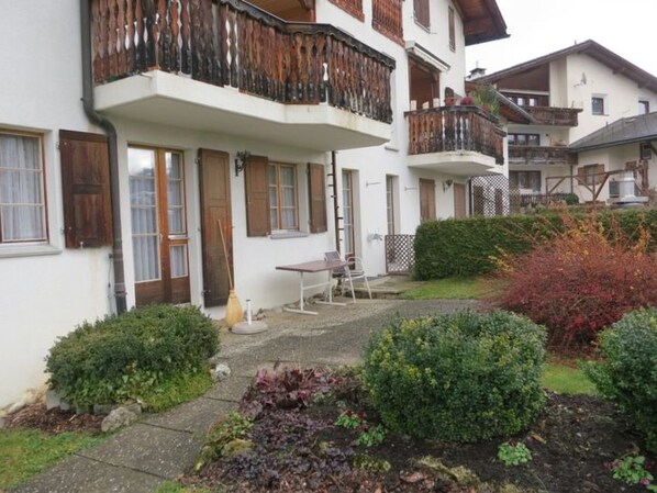 Un edificio residencial de dos plantas con un balcón, un pequeño patio con mesa y sillas, y un jardín con diversas plantas.