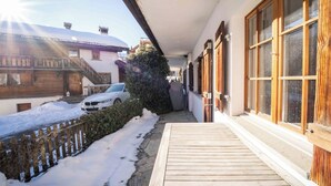 A wooden deck with a snow-covered ground, a white car parked in the driveway, and a building with wooden shutters and a balcony. - Casa Fulin, Obersaxen Affeier, (Obersaxen Affeier), 34075B by Interhome (Obersaxen Affeier)