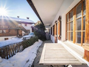 A wooden deck with a snow-covered ground, a white car parked in the driveway, and a building with wooden shutters and a balcony. - Casa Fulin, Obersaxen Affeier, (Obersaxen Affeier), 34075B by Interhome (Obersaxen Affeier)