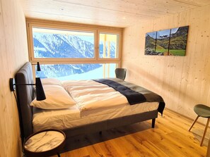 A bedroom with a bed, a chair, a round table, and a window with a mountain view. - Ferienhaus Solar by biobuchli, Tenna, (Tenna), 06007B by Interhome (Tenna)