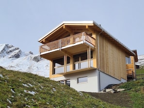 A wooden chalet with a balcony, set against a mountainous backdrop.