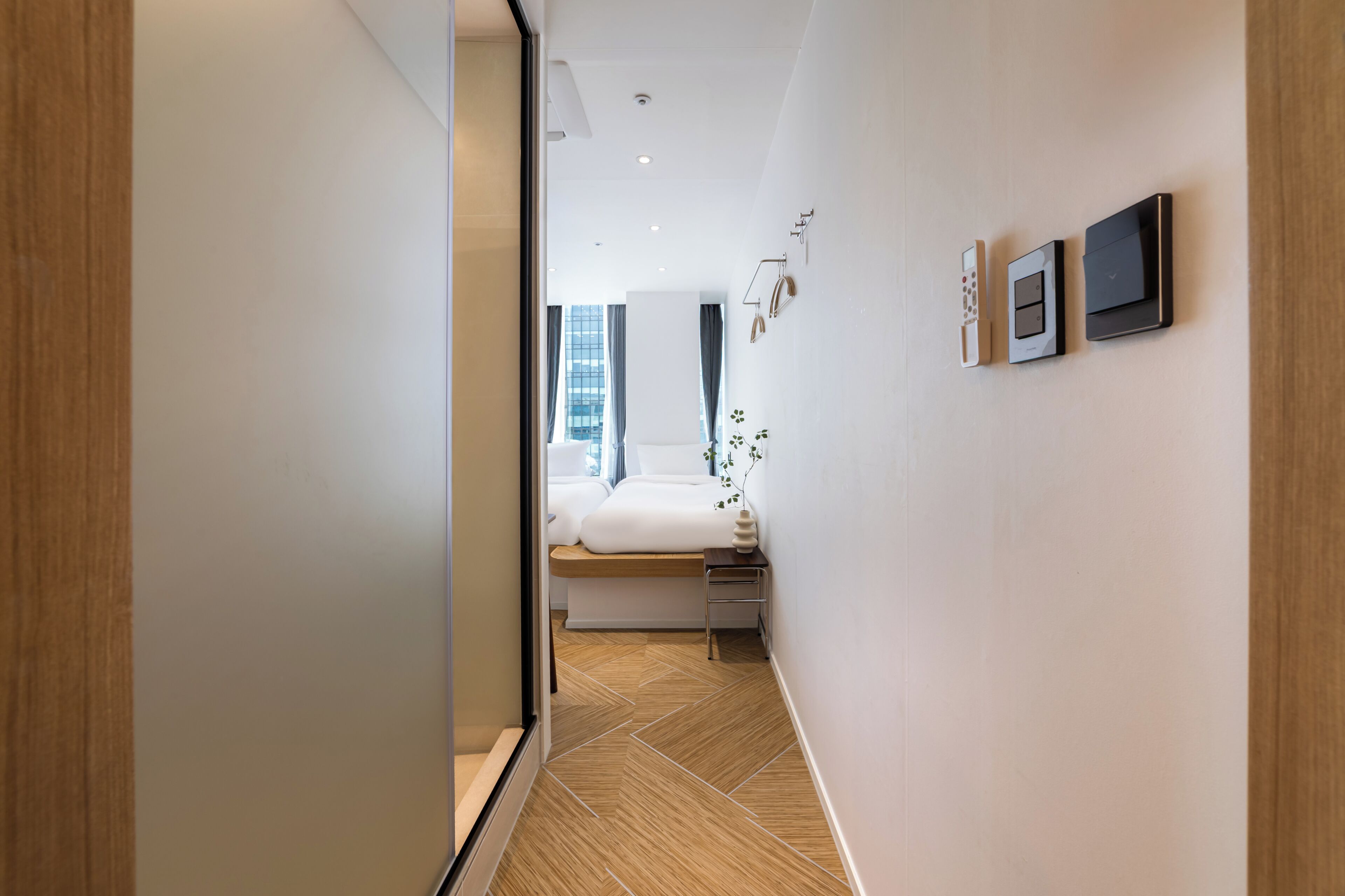 Una habitación de hotel moderna con suelo de madera, una cama con ropa de cama blanca, una mesita de noche y un panel de control en la pared.