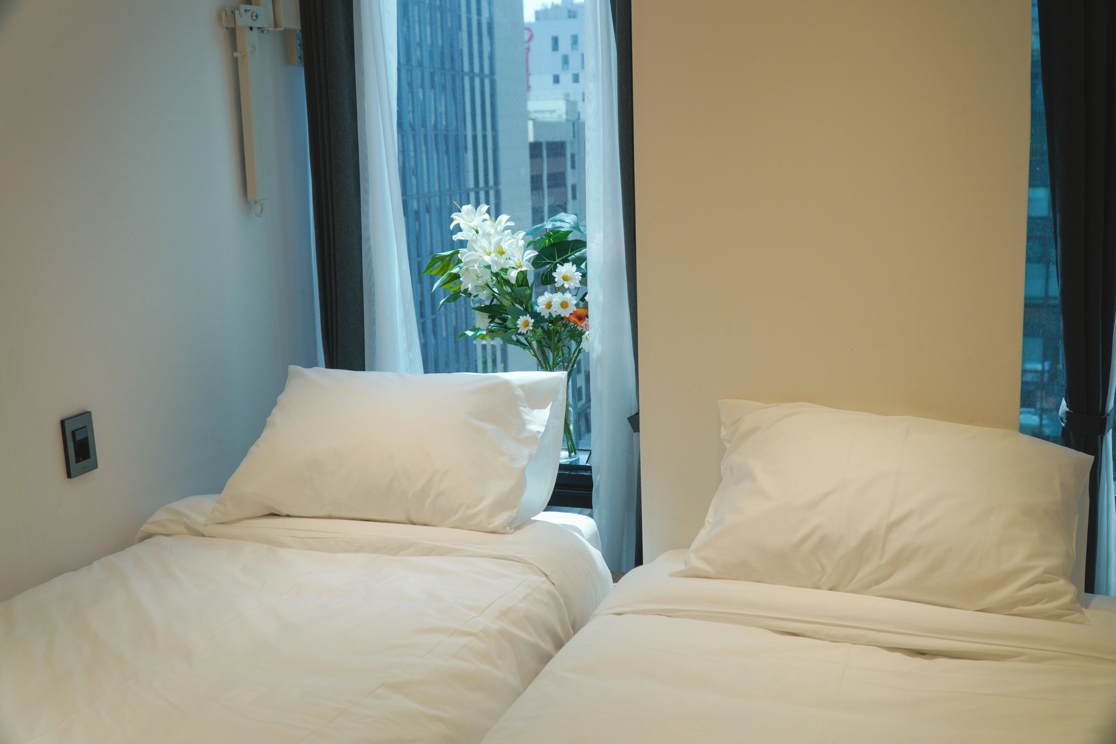 A hotel room with two beds, a window with a view of buildings, and a vase with flowers.