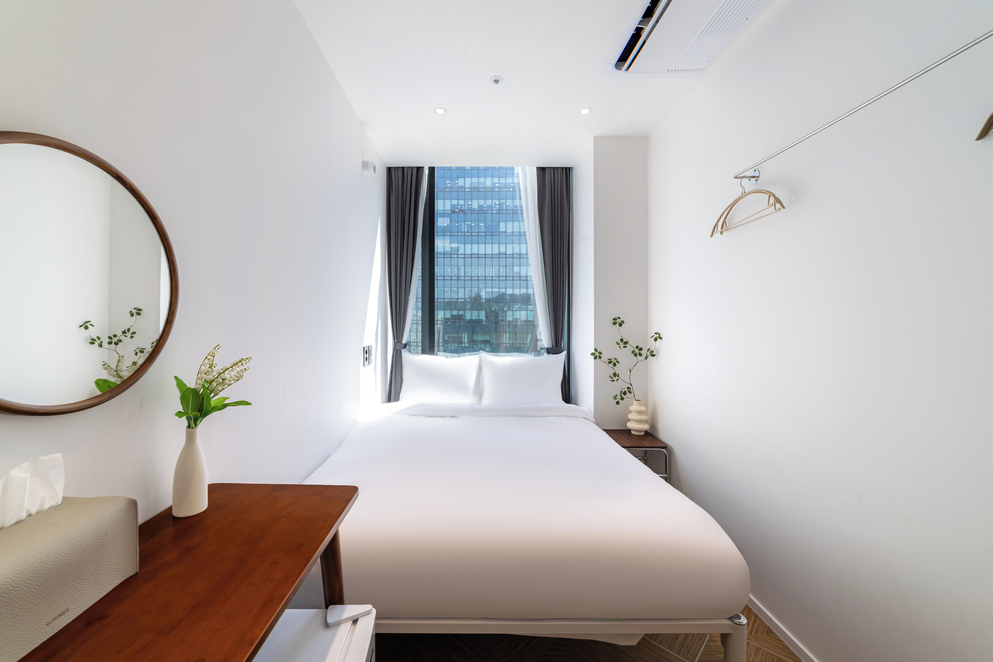Une chambre d’hôtel avec un lit, une table de chevet ornée d’un vase, un miroir, une fenêtre avec des rideaux et une vue sur un immeuble.